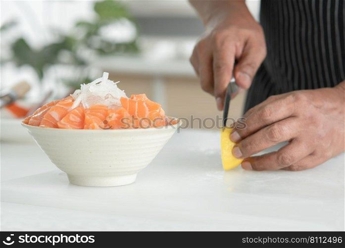 Selective focus at radish on sliced fresh salmon sashimi on bowl with ice. Asian senior man"s hand holding knife slicing lemon on cutting board on background. Japanese food home cooked concept