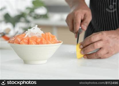 Selective focus at radish on sliced fresh salmon sashimi on bowl with ice. Asian senior man"s hand holding knife slicing lemon on cutting board on background. Japanese food home cooked concept