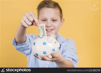 selective focus boy inserting currency note polka dot ceramic piggy bank against yellow backdrop