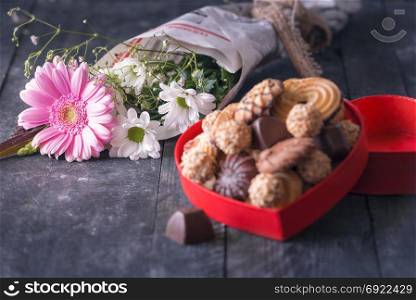 Selective focus image with a lovely bouquet of chrysanthemum flowers, wrapped in old newspaper and a blurred box full of sweets, on a black wooden background.
