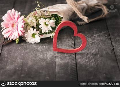 Selective focus image with a lovely bouquet of flowers wrapped in a newspaper and a red blur heart, on a black wooden background.