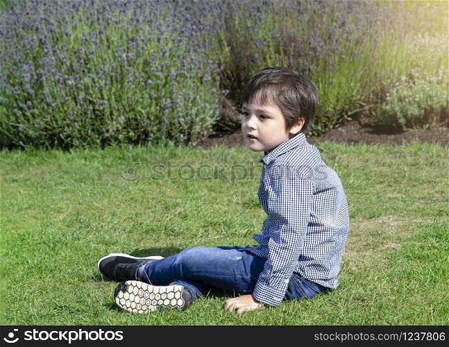 Selective focus little boy with smiling face sitting on grass in lavenders field, Portrait of happy kid playing outdoors with blurry flowers background, Child having fun in lavender garden in the summer.