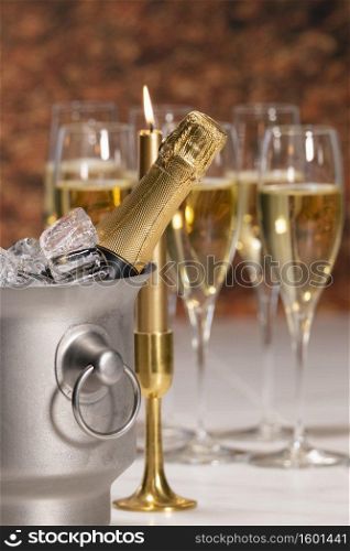 Selective focus of a champagne bucket with ice cubes and a champagne bottle with an out of focus candle and champagne glasses. Celebration concept.