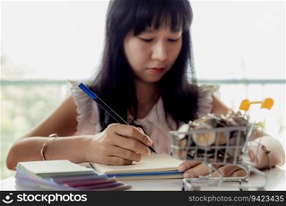 Selective focus of Asian woman’s hand writing on a notebook with blurred money and coins in a shopping cart for business and financial planning concept