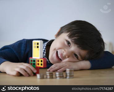 Selective focus of child boy playing plastic door and window frame with blurry foreground of stacks coins on wooden table. Learning financial responsibility and planning savings concept.