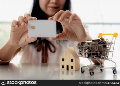 Selective focus of hand holding a blank credit chip card with wooden house model and full of coins in shopping cart for planning, savings and property investment concept