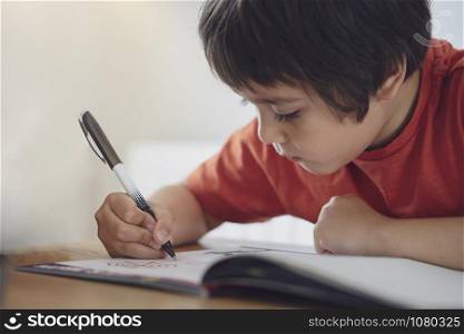 Selective focus of school kid boy siting on table doing homework, Happy Child holding black pen,Little boy is writing on white paper, Elementary school, Homeschooling concept