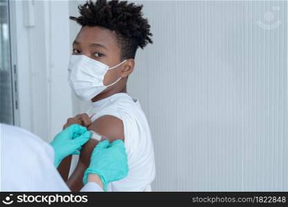 Selective focus on African American boy wear mask looking at camera while doctor's hands with gloves putting plaster on his upper arm after man get vaccinated. White background. Copy space