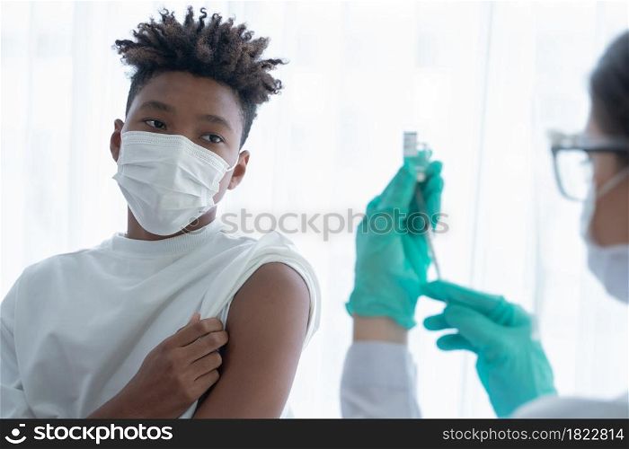 Selective focus on African American boy wear mask roll up his sleeves looking at Caucasian woman doctor wear gloves using syringe to suck vaccine or medicine out of vial. Vaccinating people concept