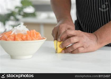 Selective focus on Asian senior man"s hand holding knife slicing lemon on cutting board for decorate sliced fresh salmon sashimi on bowl with ice and radish. Japanese food home cooked concept