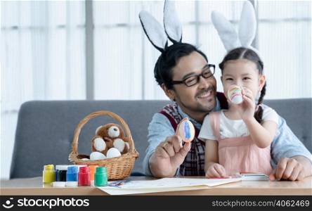 Selective focus on decorated Easter eggs in hands of Caucasian little girl daughter and young Asian father in bunny ears headbands in living room at home. Happy family prepare for Easter holiday