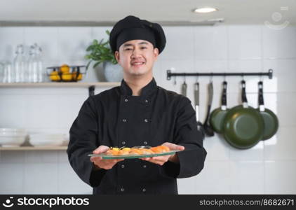 Selective focus on delicious sushi on plate in hand of young handsome Asian chef with black uniform and hat, Japanese food, in restaurant kitchen