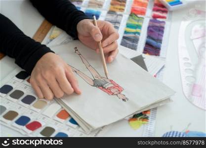 Selective focus on fashion designer hand, holding pencil, sketching creative clothes on paper, with fabric and threads color chart, tailor tools on working desk at workplace