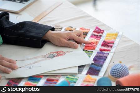 Selective focus on hands of fashion designer comparing or choosing color of threads with color of fashion clothes on sketch paper, with fabric and threads color chart and tailor tools on working desk