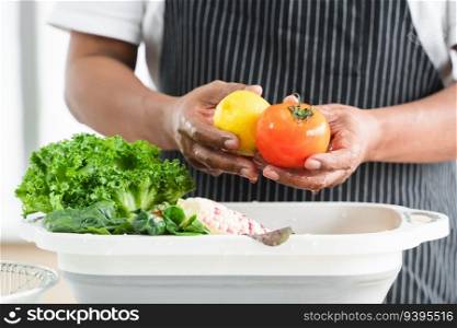 Selective focus on hands of senior man washing tomato and lemon at home. Vegetables, lettuce and corn soak with water in basin. Healthy food and cleaning before cooking concept. Focus on tomato