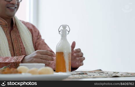 Selective focus on homemade fermented raw drink kombucha tea bottle, gas gushing out of the bottle. Happy Indian man open bottle cap wearing traditional clothes, sitting at dining table. Copy space