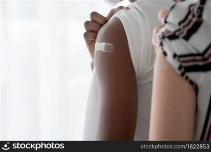 Selective focus on medicated plaster stuck on upper arm of dark skinned young man wear mask rolling up sleeves to show that he get vaccinated. Caucasian woman on blur foreground. White background