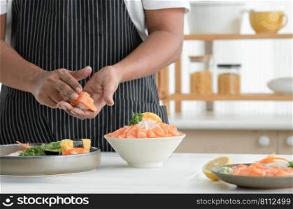 Selective focus on old man chef hand making salmon sushi and prepare sliced salmon sashimi on ice in bowl served with wasabi, sliced lemon and radish at kitchen. Japanese food home cooked concept