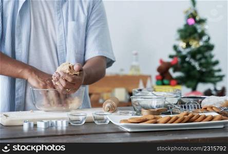 Selective focus on senior man hands kneading gingerbread cookie dough in mixing bowl on table with brown cookies in plate, rolling pin, mold and flour at home kitchen decorated with Christmas tree