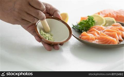 Selective focus on senior man’s hand grinding fresh wasabi in a small bowl with sliced fresh salmon sashimi in decorated plate ready to serve. Japanese food home cooked concept