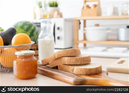 Selective focus on sliced whole wheat bread on wooden tray with apricot jam in jar, bottle of fresh milk, oranges, avocado and watermelon for juices. Breakfast ingredients are on kitchen table at home