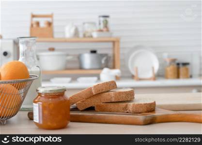 Selective focus on sliced whole wheat bread on wooden tray with apricot jam in jar, bottle of fresh milk, oranges in basket for juices. Breakfast ingredients are on kitchen table at home