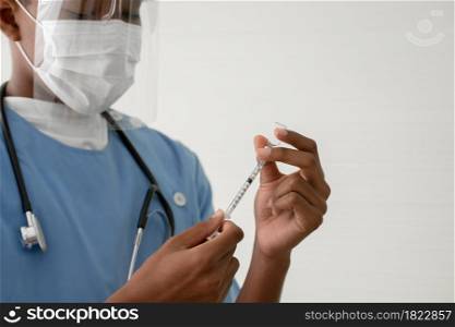 Selective focus on Young African American man doctor's hand using syringe to suck vaccines or liquid medicine out of vial bottle with mask, face shield and stethoscope at hospital
