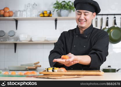 Selective focus on young handsome Asian chef with black uniform and hat, preparing delicious fresh salmon sushi, Japanese food, at restaurant kitchen