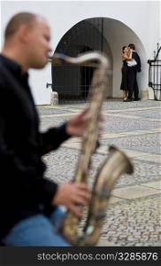 Selective focus shot of a street musician playing his saxophone while a romantic couple kiss in the background