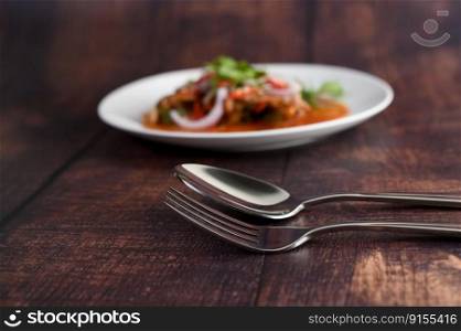 Selective focus stainless spoon and fork on wooden table, blured background with Spicy salad of sardine with tomato sauce in white dish, copy space