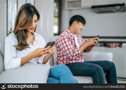 Selective focus, Young Asian Couple sitting on sofa in living room, They are enjoy with social media networks addicts surf the internet on mobile phones ignoring each other, family addicted smartphone concept