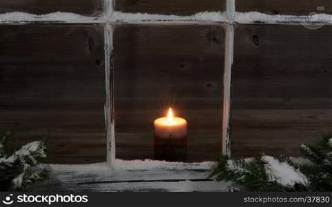 Selective view of a glowing candle through a snow covered window with fir branches.