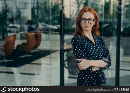 Self assured female director with red hair pleasant appearance dressed in elegant dress spectacles poses near office building waits for partner going to have meeting. Corporate worker outdoor. Self assured female director with red hair poses near office building