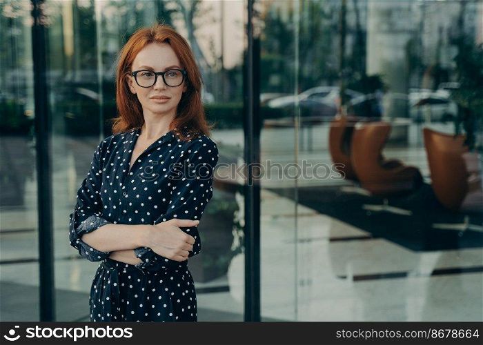 Self assured female director with red hair pleasant appearance dressed in elegant dress spectacles poses near office building waits for partner going to have meeting. Corporate worker outdoor. Self assured female director with red hair poses near office building