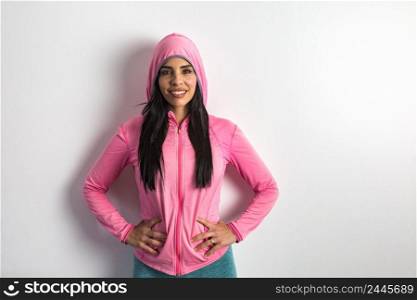 Self assured Hispanic female athlete in pink hoodie holding hands on waist and looking at camera with smile while standing against gray wall before fitness training. Confident Hispanic sportswoman ready for workout