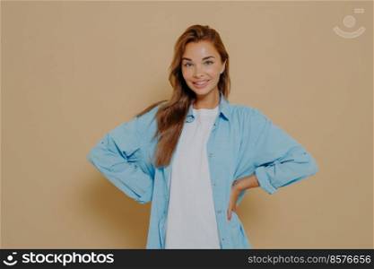 Self assured smiling lovely model keeps hands on waist, wears long blue shirt over white tshirt, looks directly at camera, expresses her right, isolated over beige background. Self confidence concept. Image of self assured smiling lovely model keeps hands on waist