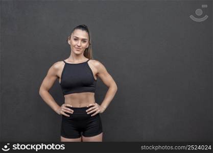 Self assured strong sportswoman in black shorts and crop top holding hands on waist and looking at camera while standing against black wall during training in gym. Confident female athlete with hands on waist