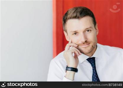 Self assured unshaven male office worker in formal white shirt with tie, holds hand on cheek, thinks about starting new stage in life, poses against red and white background. Business concept