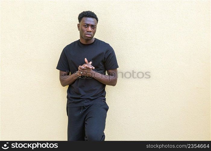 Self assured young African American male with dark hair in black casual wear standing near colored concrete wall and looking at camera. Unemotional young black guy standing near wall on street