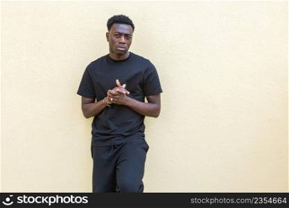 Self assured young African American male with dark hair in black casual wear standing near colored concrete wall and looking at camera. Unemotional young black guy standing near wall on street