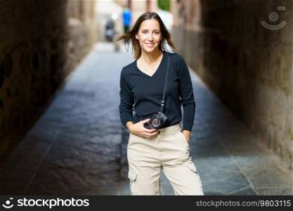 Self assured young female photographer with camera in casual clothes standing on narrow street with hand in pocket and looking at camera during vacation. Smiling woman with photo camera looking at camera on narrow street