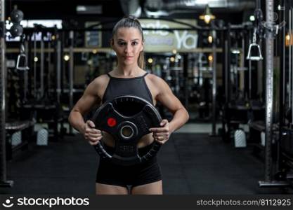 Self assured young muscular female athlete with ponytail in sportswear holding heavy weight plate and looking at camera while exercising in modern gym. Strong lady exercising with weights in contemporary sports club
