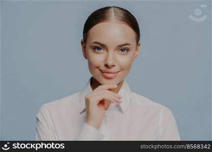 Self confident attractive businesswoman holds chin and looks directly at camera, wears elegant white shirt, has healthy skin and natural makeup, isolated over blue background. Women, business, career