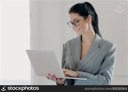 Self confident cheerful woman economist analyzes report about company income on laptop computer, shares multimedia files, has dark hair combed in pony tail, dressed in formal outfit, stands indoor