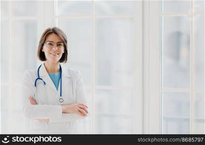 Self confident professional doctor stands with arms crossed, wears white medical gown with stethoscope, thinks about work positively, poses against big window. Healthcare and occupation concept