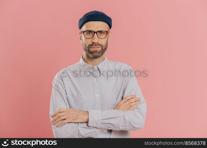 Self confident unshaven man crosses hands over chest, wears spectacles and white shirt, isolated over pink background. Bearded hipster ready for business meeting. People and fashion concept.