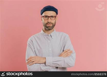 Self confident unshaven man crosses hands over chest, wears spectacles and white shirt, isolated over pink background. Bearded hipster ready for business meeting. People and fashion concept.