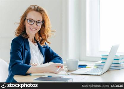 Self confident young female director plans work process, writes main theses in notepad, rewrites information from laptop computer, drinks tea, poses over office interior. Technology, occupation