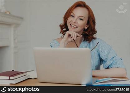 Self employed female freelancer has gentle smile on face, works on startup project, poses with laptop computer at desktop, searches information for productive work, sits indoors, holds eyewear