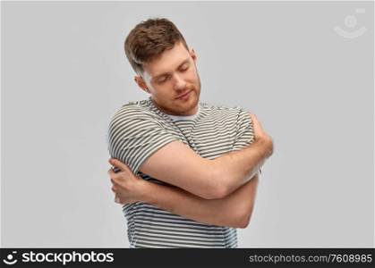 self loving, care and people concept - happy young man in striped t-shirt hugging himself over grey background. happy young man in striped t-shirt hugging himself